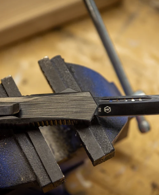 A close-up of a black folding knife held in a blue bench vise against a wooden workbench background. The knife has a textured black finish and a small white logo with the letter 'B' on the blade.