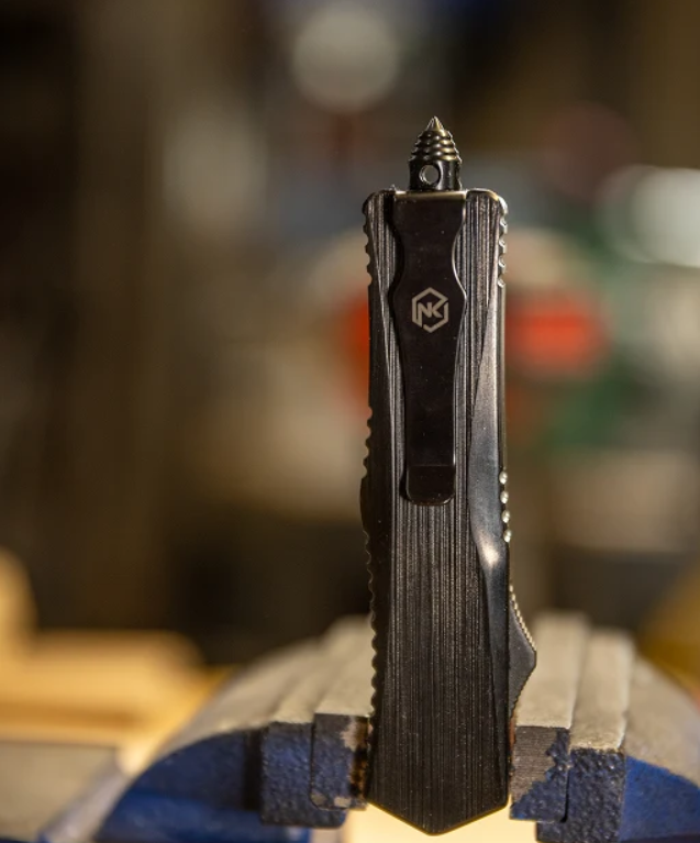 A black pocket knife with a textured handle and a visible MK logo, shown standing upright in a blue bench vise with a blurred workshop background.