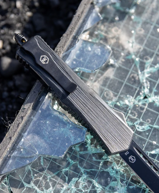 A black tactical pocket knife with a textured handle and a silver geometric logo, shown resting on broken glass over a gridded surface.