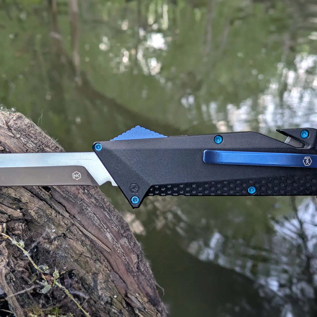 OTF D2 tanto blade knife with black anodized black handle with blue accents on a tree branch by a body of water.