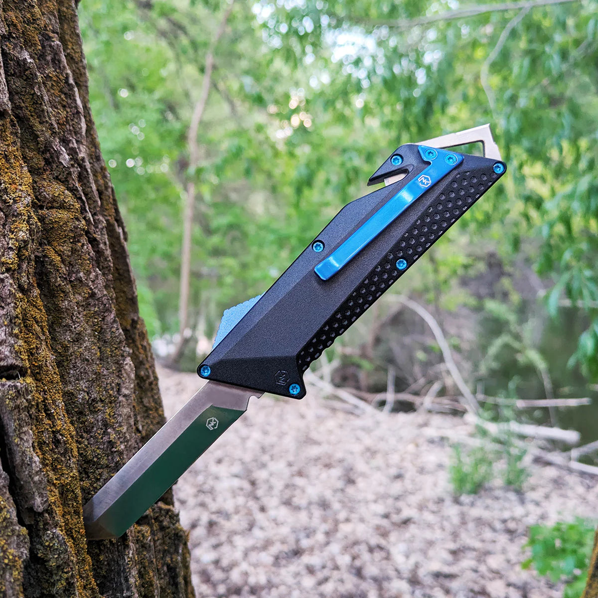 OTF knife with black anodized aluminum handle with blue belt clip, deployment switch, and screws embedded into tree trunk in the woods.