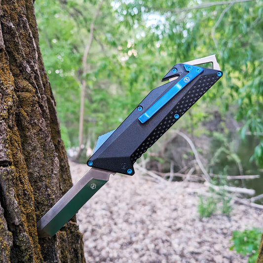 OTF knife with black anodized aluminum handle with blue belt clip, deployment switch, and screws embedded into tree trunk in the woods.